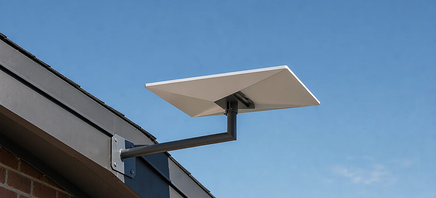 A Starlink-style flat rectangular phased-array panel carried on a black steel arm bracket fixed to the fascia board at the edge of a roof, aimed at open sky above a pine treeline, with the roof covering untouched